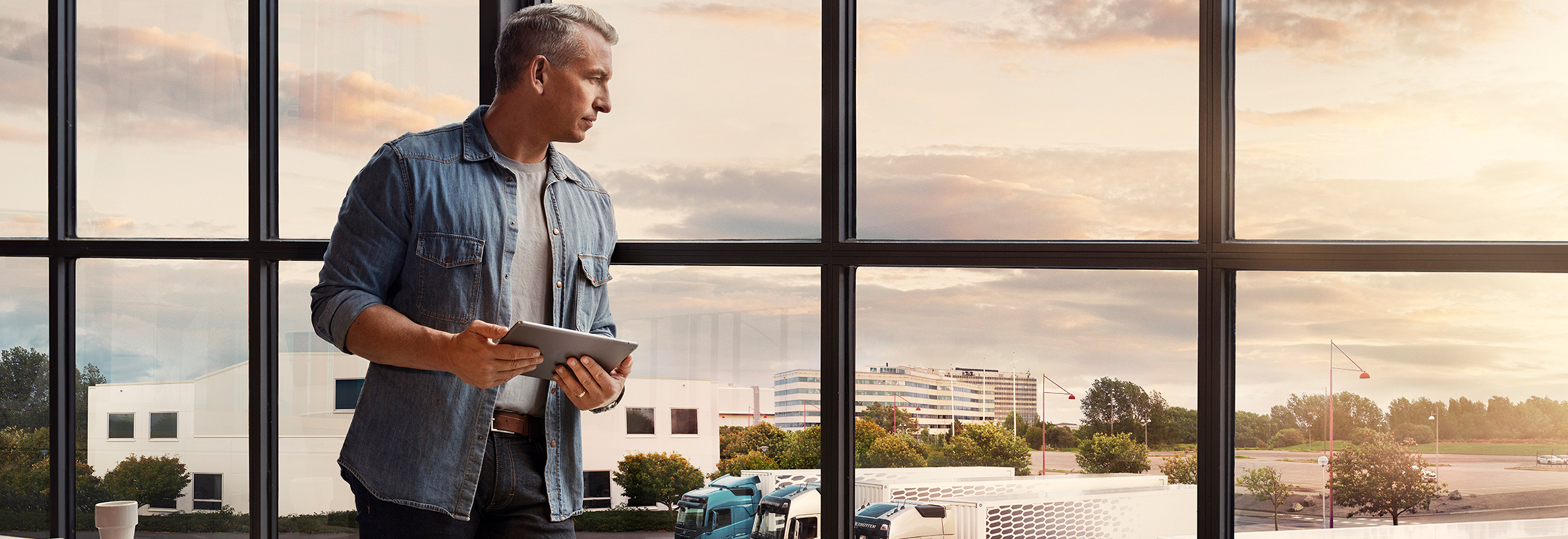En mann som holder et nettbrett, står ved et vindu og ser ned mot lastebilparken sin