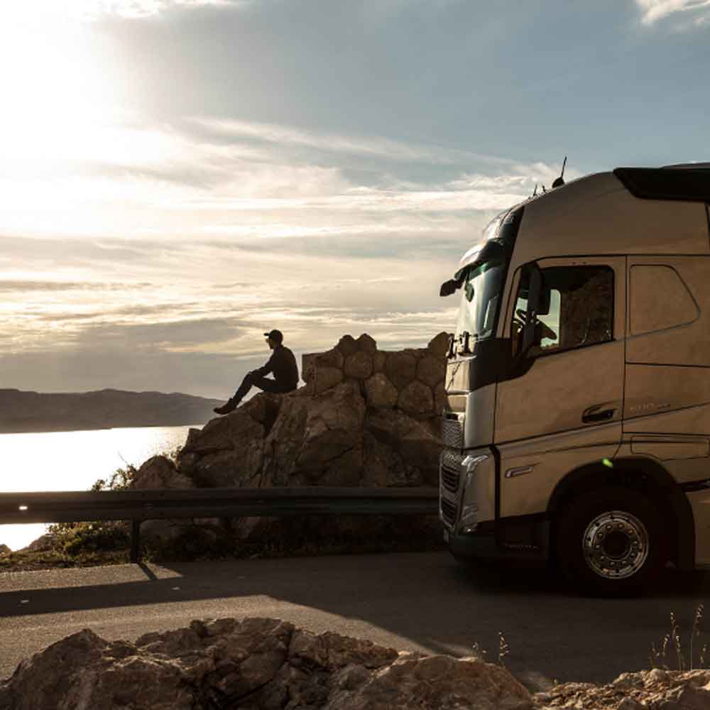 En sjåfør sitter på en stein og ser ut mot havet ved solnedgang, med sin Volvo FH-lastebil parkert ved siden av seg